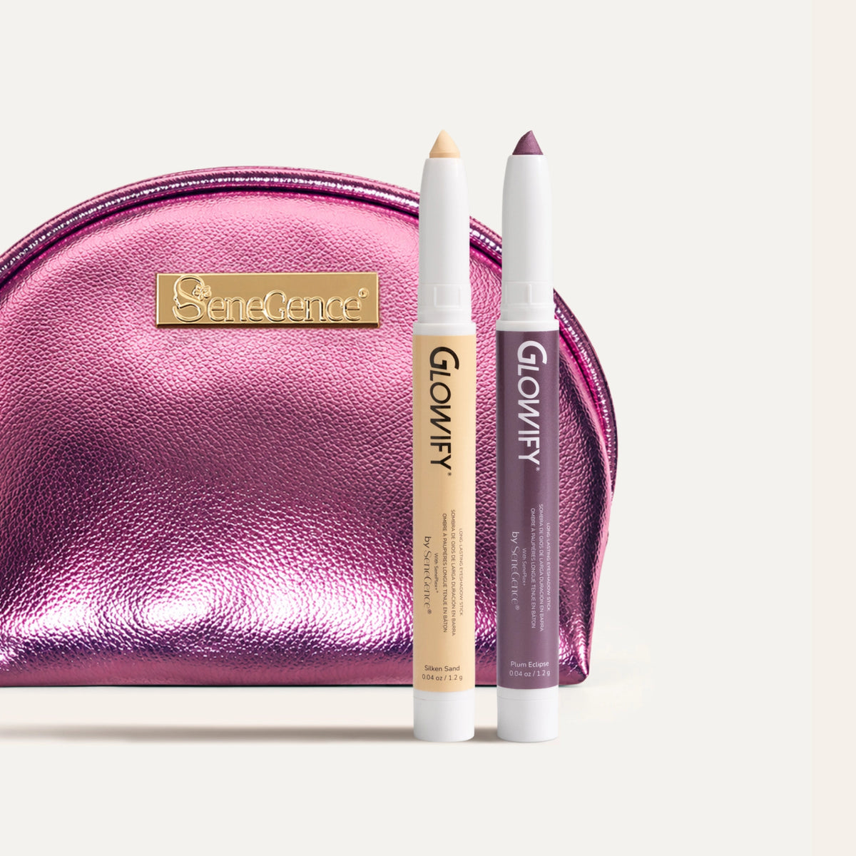 Two white-tipped eyeshadow sticks with their color tips exposed, one pale sand and one plum, standing in front of a metallic pink zip pouch with a gold SeneGence plate, Desert Dusk Glowify® Eyeshadow Stick Duo.