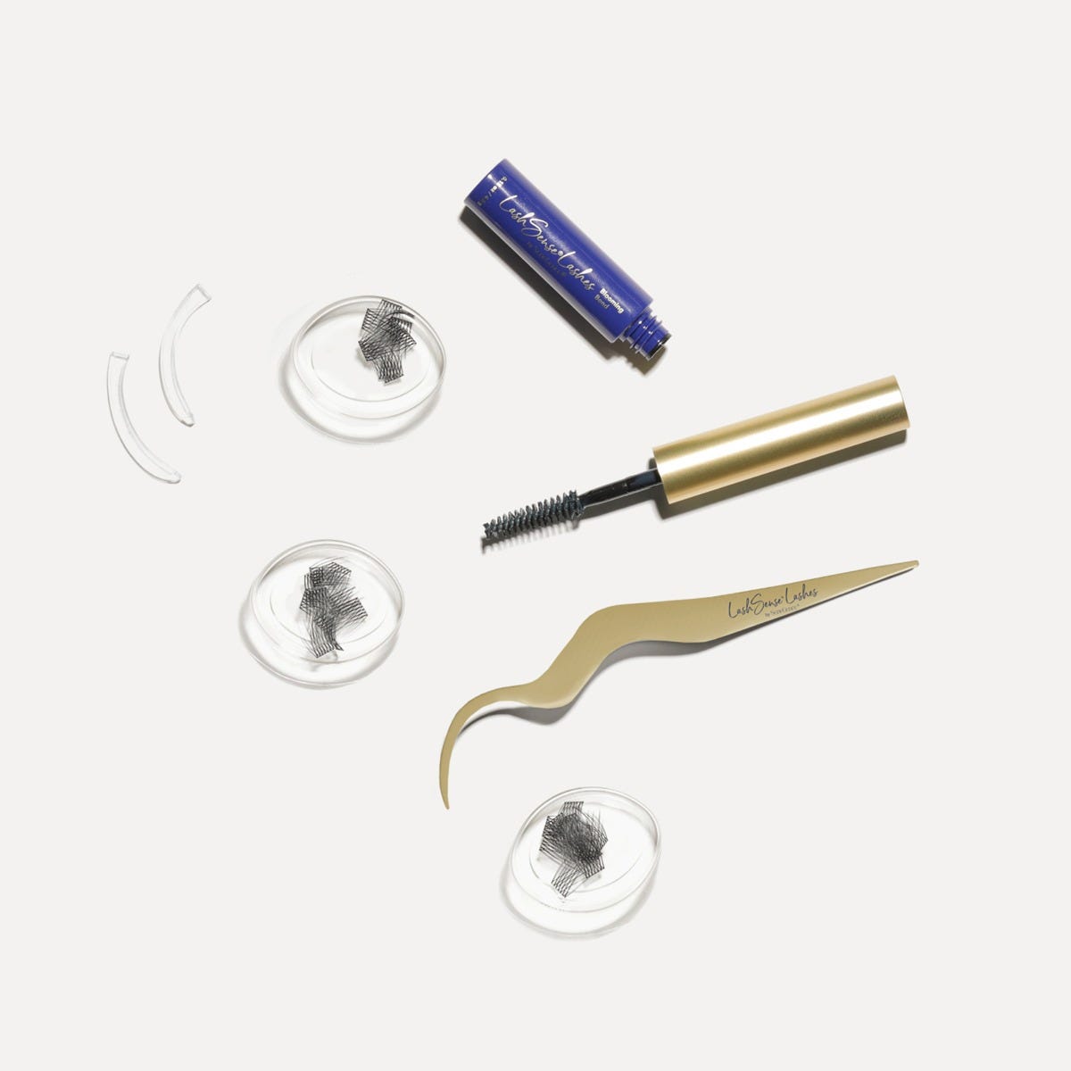 Black lash clusters in three clear round trays beside a blue adhesive tube, a gold-capped spoolie brush, two clear curved applicator guides and curved gold tweezers, LashSense® Lashes Bloom Box.