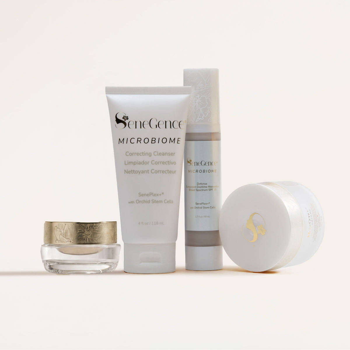A small clear glass jar with a gold orchid-patterned lid, a white Correcting Cleanser tube, a Defense Daytime Moisturizer SPF 15 pump bottle with a frosted orchid cap and a large white jar lying on its side with a gold logo lid, Microbiome SkinCare Regimen Set.